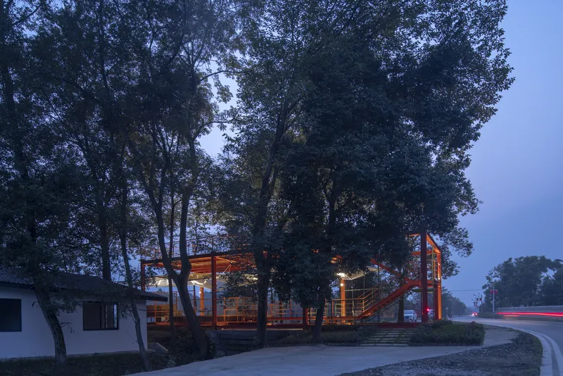 Dusk view from the road: orange steel frame glowing through the trees, stone steps, old farmhouses flanking, car light trail on the road