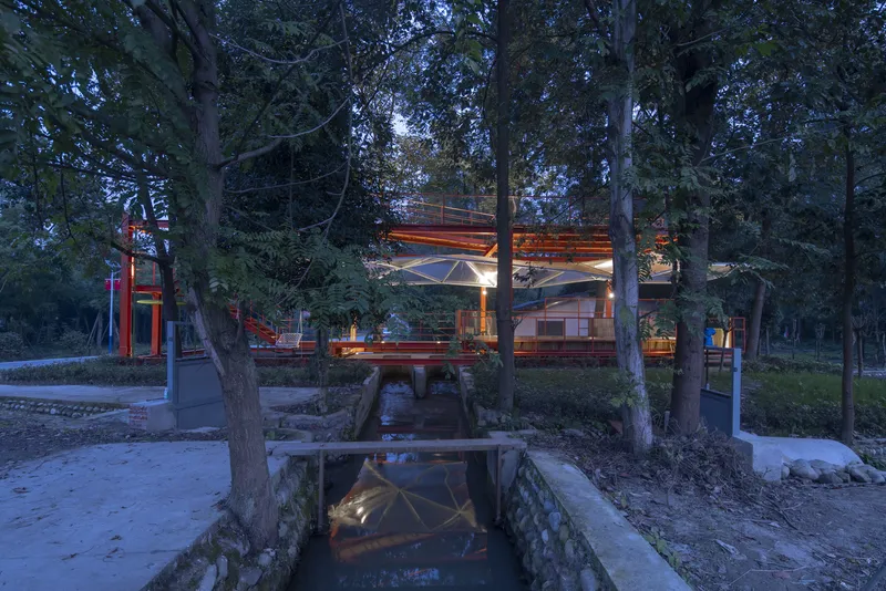Water channel approach at dusk: stone-lined channel with small bridge, orange pavilion glowing beyond, tall trees on both sides, gravel path