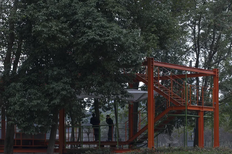 Two figures on the upper viewing platform, orange steel stair and railing, trees towering above, overcast sky