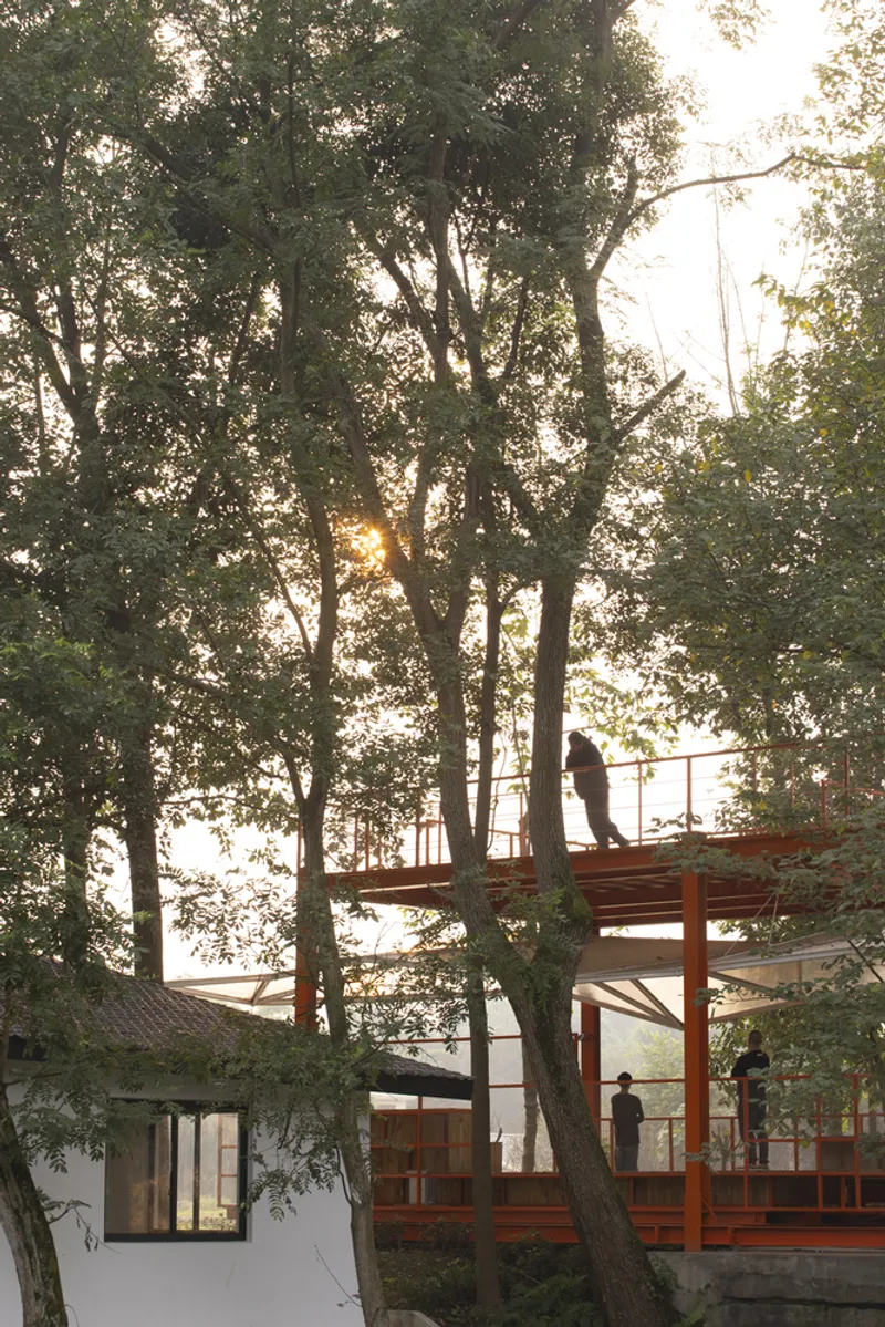 Orange pavilion framed by tree trunks, upper viewing deck visible, old white mill building behind, warm afternoon light