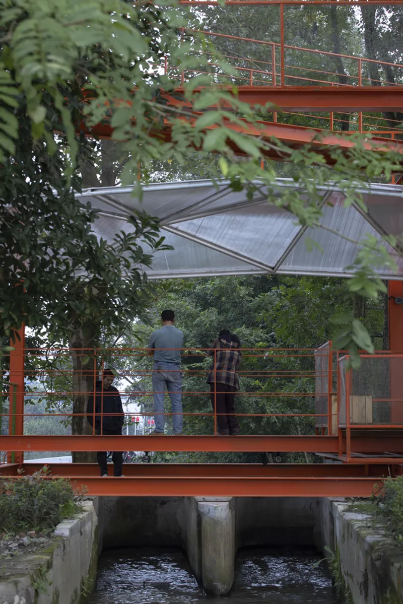 Two figures on the bridge over the water channel, orange pavilion structure above, translucent canopy, dense tree canopy overhead