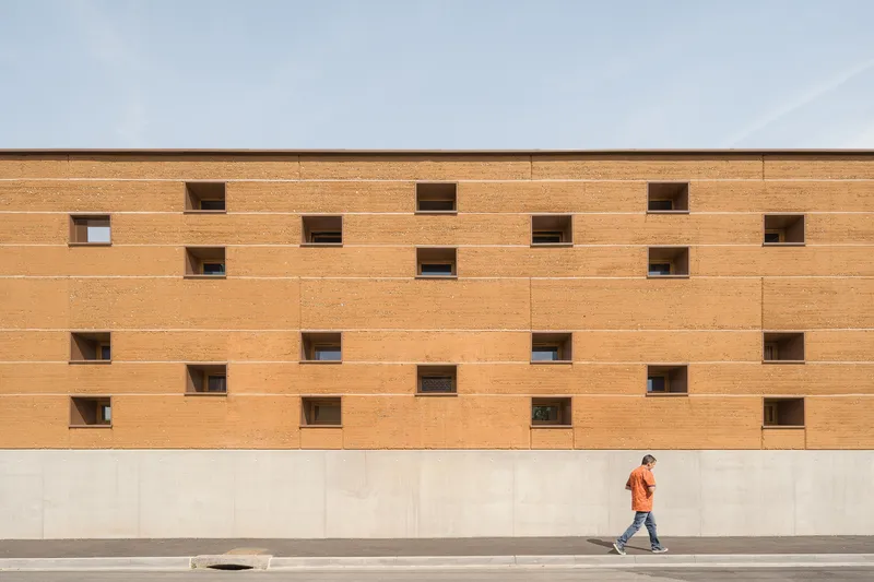 Street facade: long rammed earth wall with scattered small square windows, figure walking past