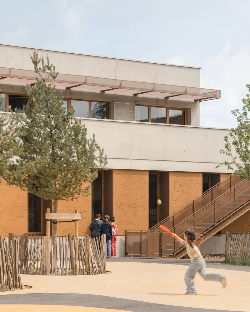 Corner view: rammed earth upper block above concrete ground floor, external steel stair, child playing with frisbee, timber fence
