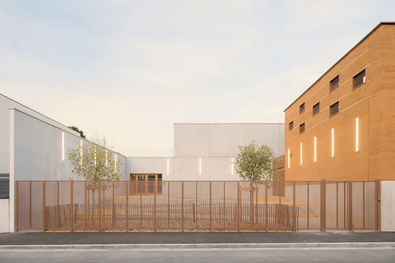 Street view: white concrete gymnasium at left, rammed earth classroom block at right, timber fence, young olive trees