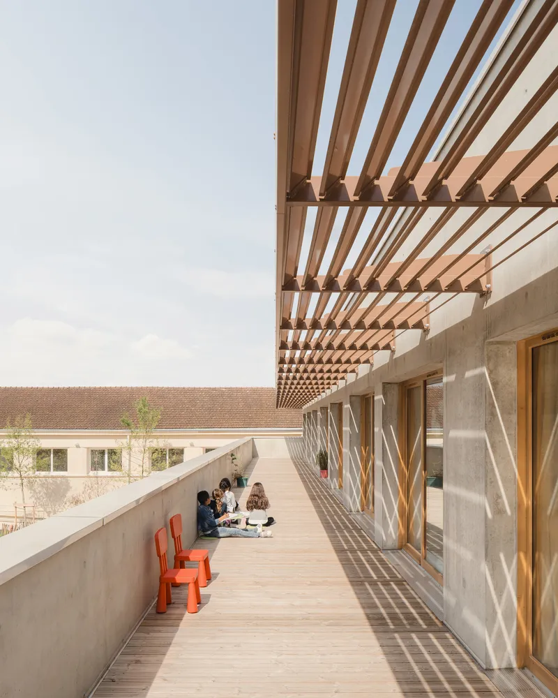Upper walkway: timber louvre pergola providing shade, concrete parapet, students sitting, red chairs, rammed earth wall behind