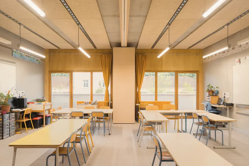 Classroom: plywood wall panels, timber-framed windows with yellow curtains, CLT timber ceiling panels, student desks and coloured chairs