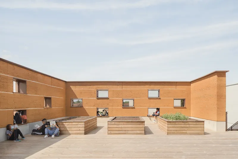 Rooftop courtyard: rammed earth walls with scattered windows, timber bench planters, students sitting and talking