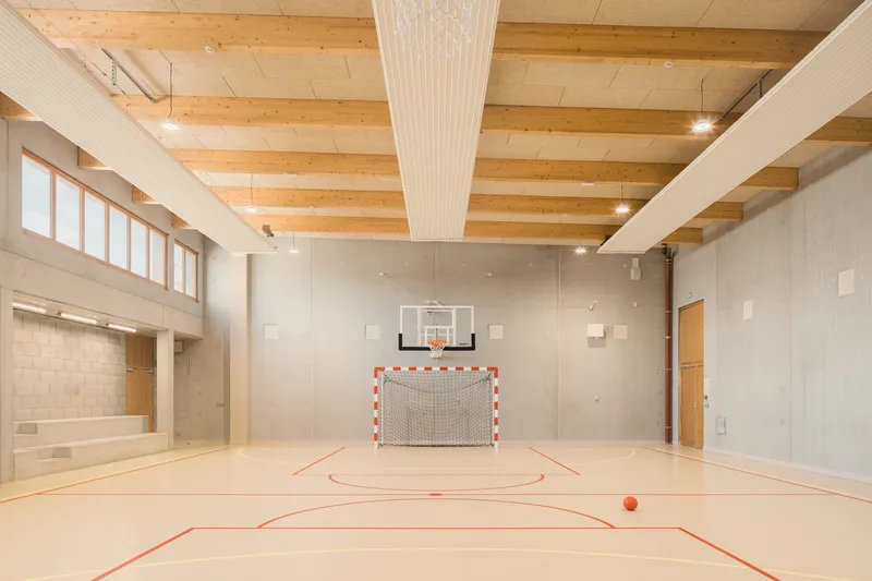 Gymnasium: glulam timber beam ceiling, basketball hoop, handball goal, timber sports floor, concrete walls, clerestory windows