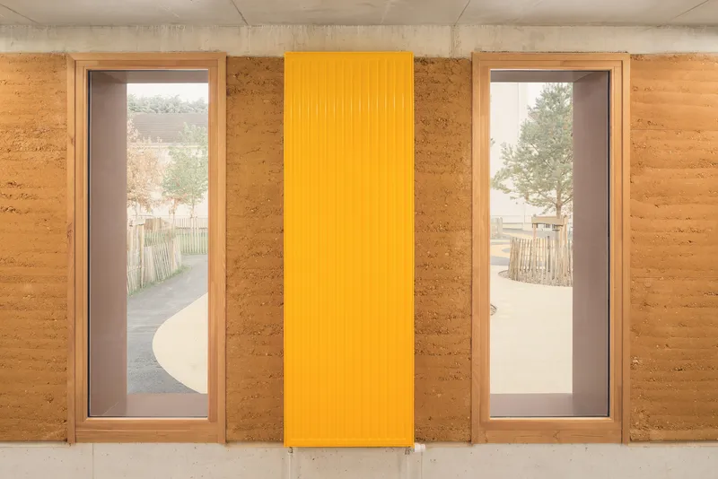 Detail: rammed earth wall with two windows flanking a yellow corrugated metal panel, courtyard visible through glass