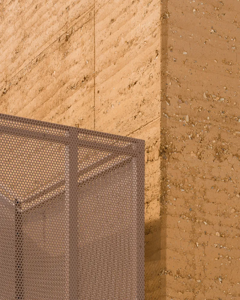 Detail: rammed earth wall corner meeting perforated metal mesh screen, warm afternoon sunlight on the earth texture