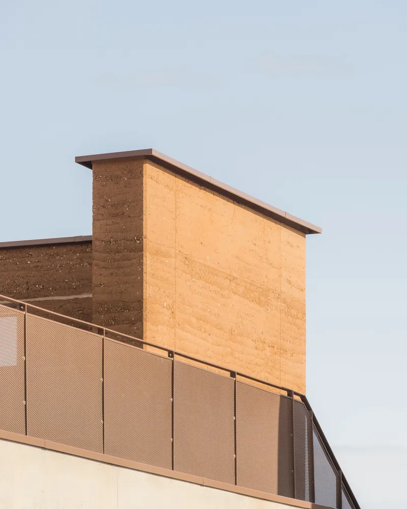 Upper level: rammed earth wall with small windows, metal balustrade walkway, concrete pilotis below