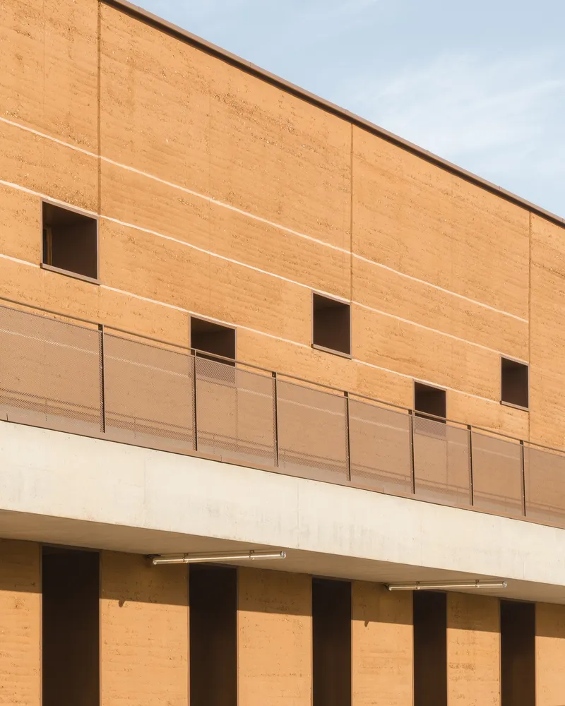 Facade: rammed earth upper floor with metal panel balustrade, concrete pilotis at ground level creating covered play area