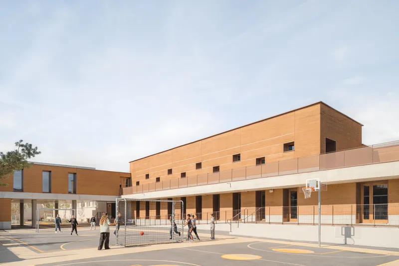 Playground: two-storey rammed earth block, covered ground floor, children playing on the asphalt, metal fence