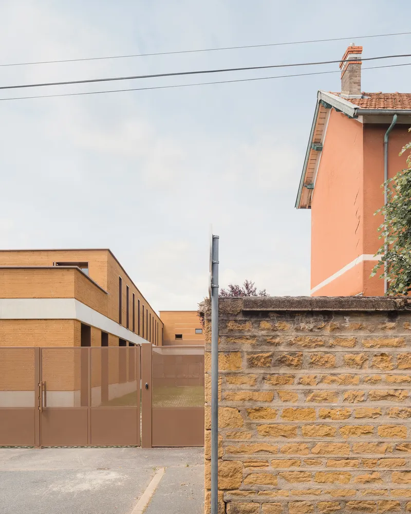 Context: rammed earth school beside an older pink rendered house, stone boundary wall, overhead wire, neighbourhood scale