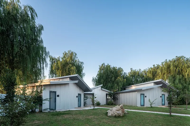 Cluster of corrugated metal pavilions with sloped roofs amid willow trees and grass