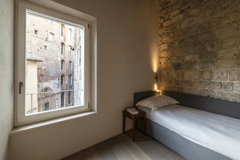 Single bed against exposed stone wall with window framing adjacent masonry buildings