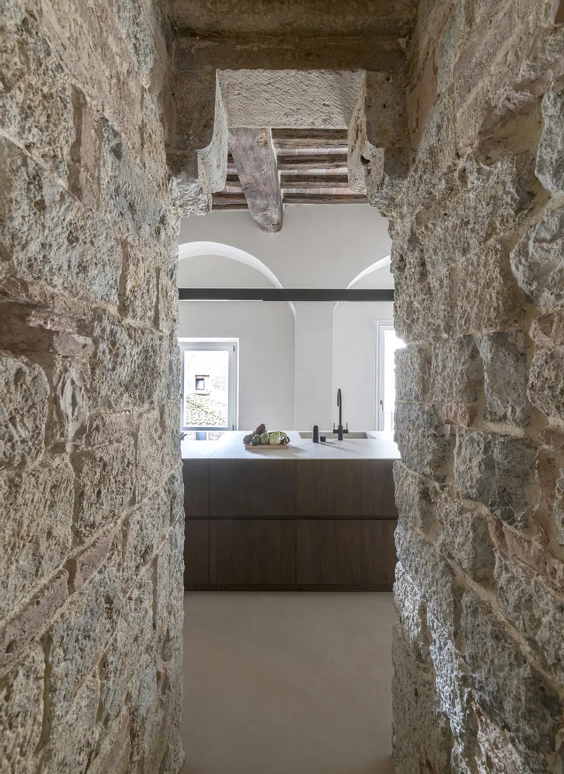 View through exposed stone doorway into kitchen with white countertop and timber cabinetry under vaulted ceiling