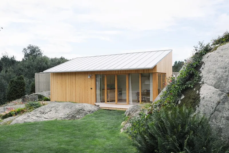 Timber and metal-roofed structure on sloping terrain between exposed rock formations and grass lawn