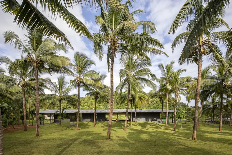 Low pavilion with overhanging roof set among coconut palms on a mowed lawn under partly cloudy skies