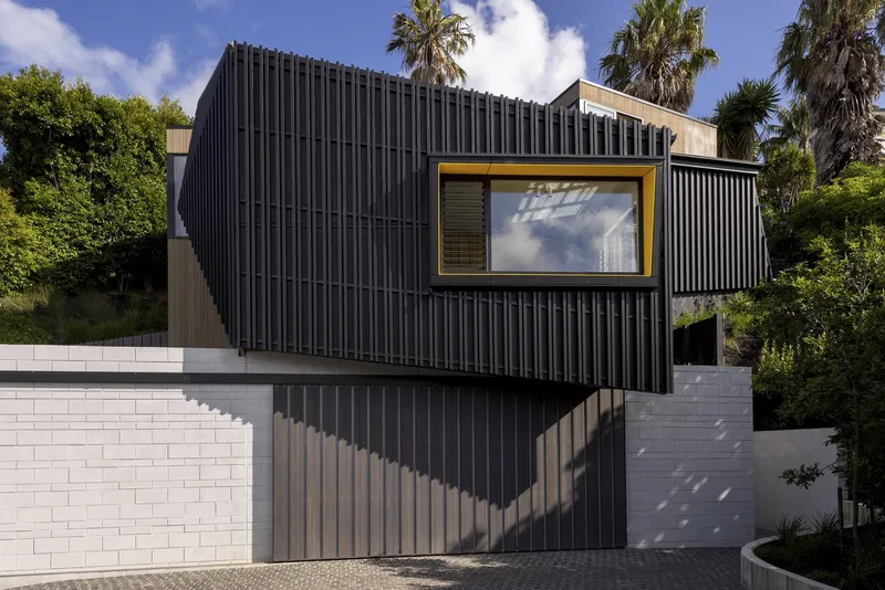 Vertical corrugated metal facade with yellow-framed window above white block retaining wall and palm trees