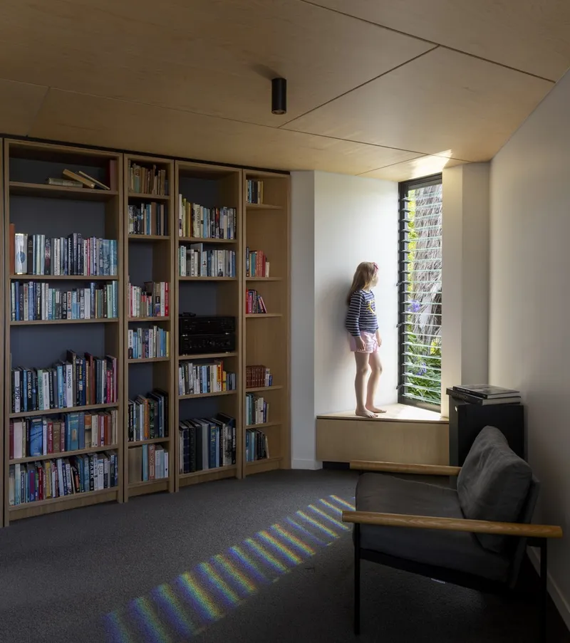 Library with timber shelving and window seat where a child stands silhouetted against louvered glass