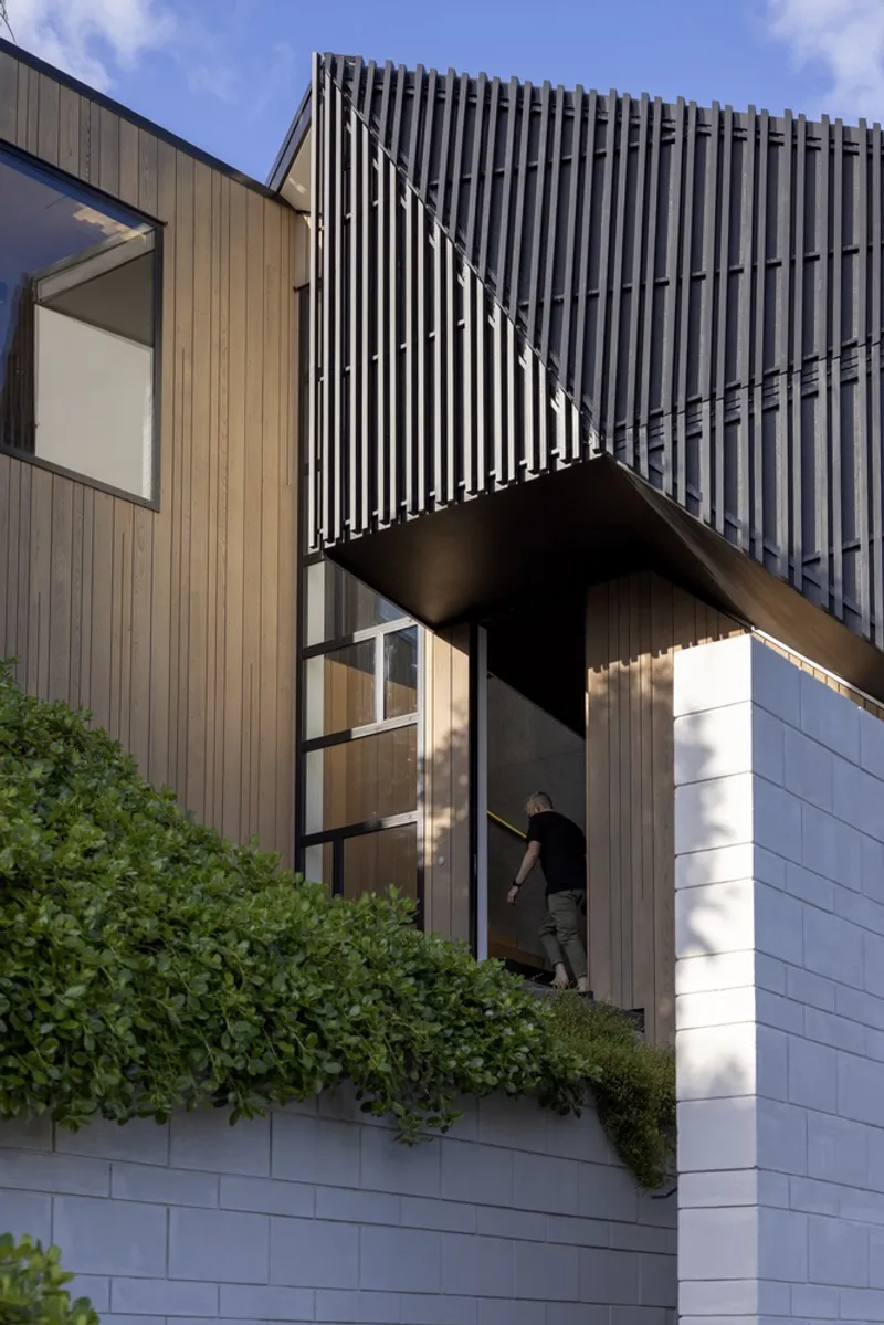 Cantilevered upper volume with vertical metal screening above timber-clad bridge and white tiled base