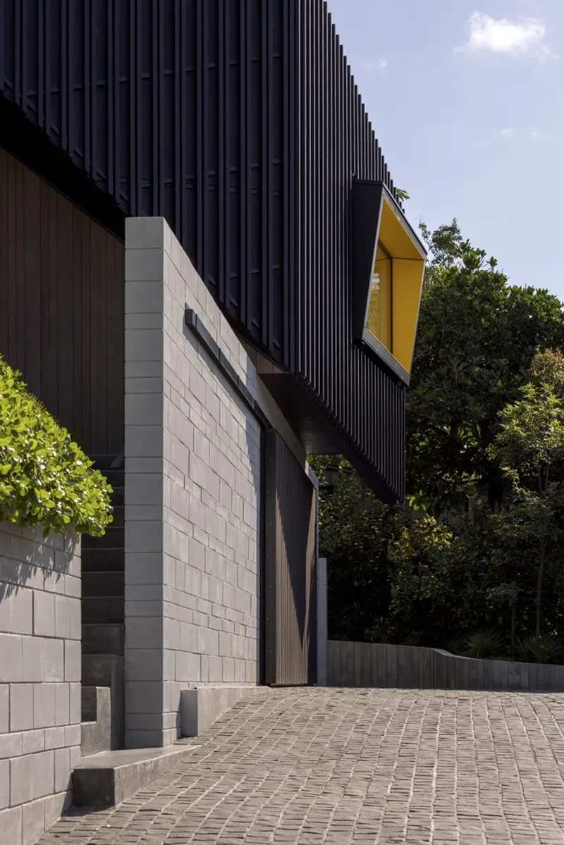 Entry courtyard showing corrugated metal volumes, yellow window frame, and concrete block walls meeting cobbled paving