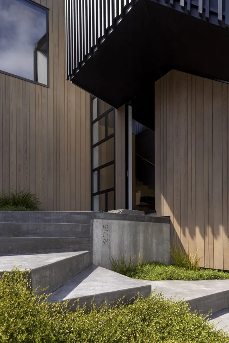 Entry sequence with concrete steps and black steel framed doorway beneath a cantilevered upper volume