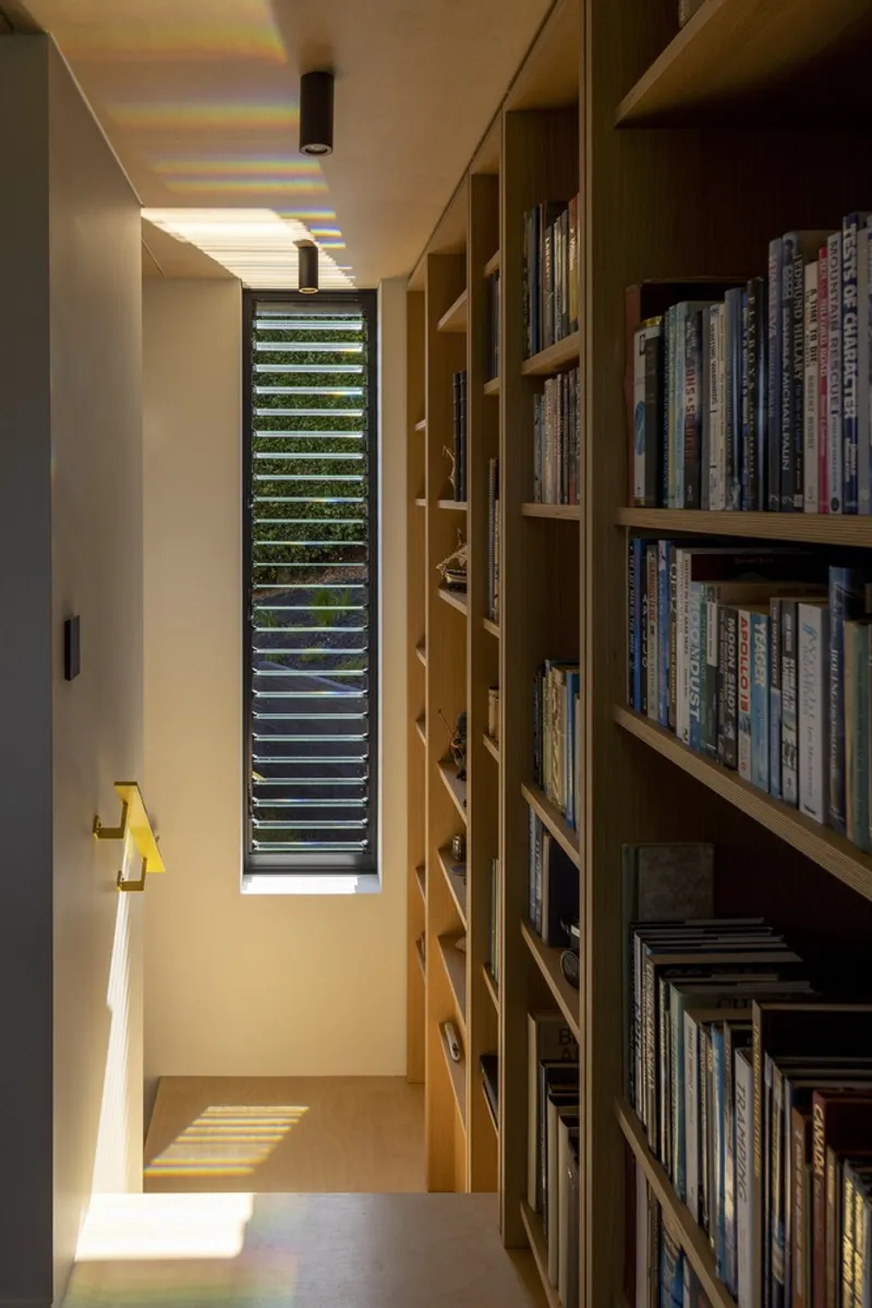 Narrow corridor with timber bookshelves and a vertical louvered window casting striped sunlight across the floor