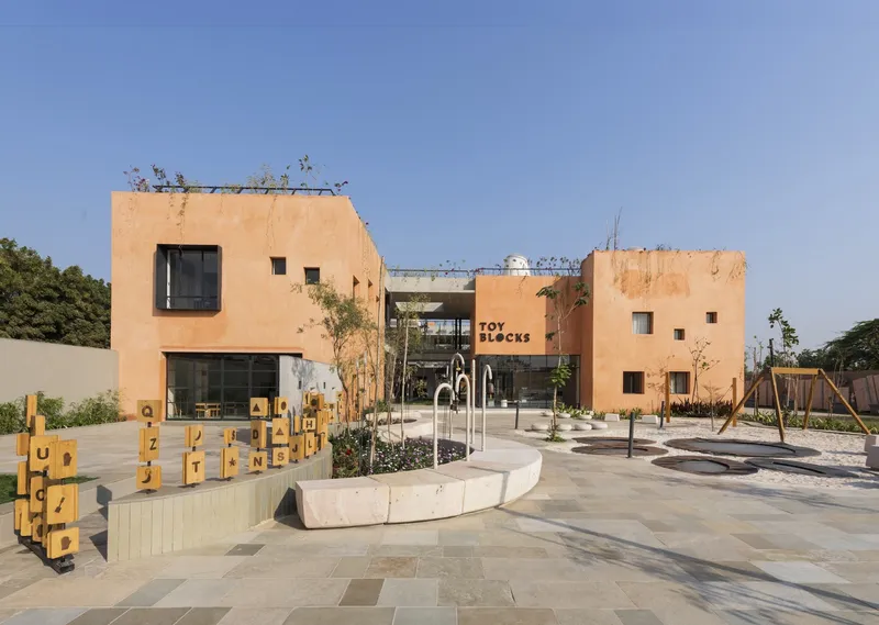 Entry courtyard with terracotta volumes, concrete planters and play equipment under afternoon sun