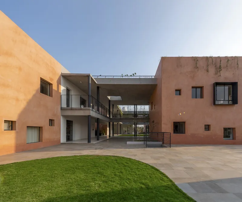 Courtyard view with terracotta-plastered volumes surrounding a paved plaza with a circular lawn and elevated walkway connecting the wings