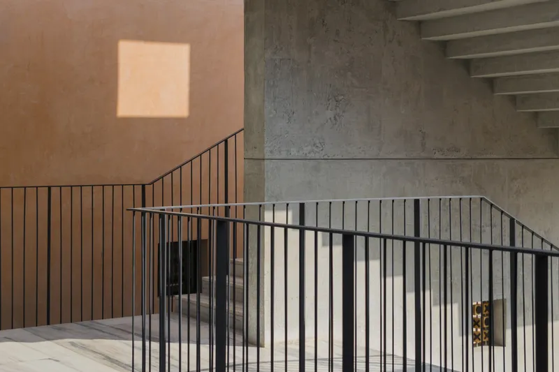 Stairwell with black metal railings set between terracotta plaster and exposed concrete walls with afternoon sunlight