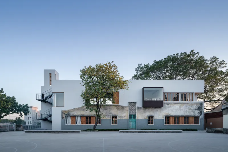 Street facade showing white volumes and concrete base with preserved trees at dusk