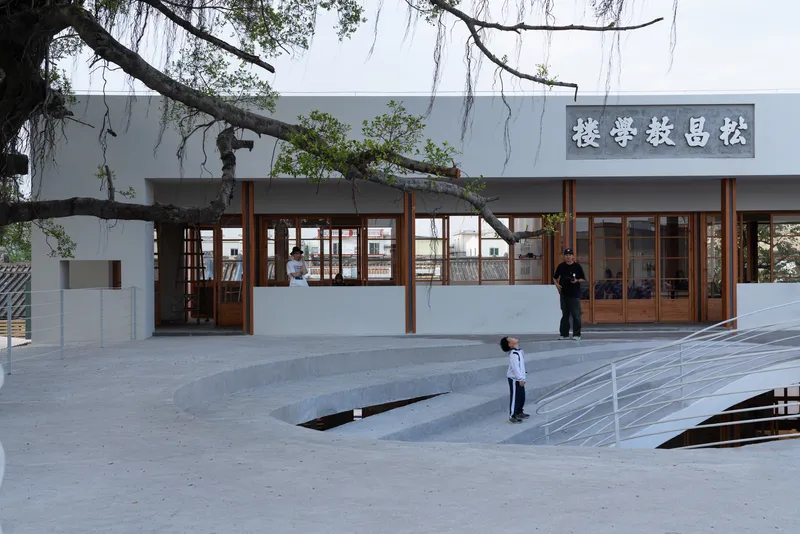Plaza with sunken courtyard opening and white facade with timber-framed glazing under willow branches