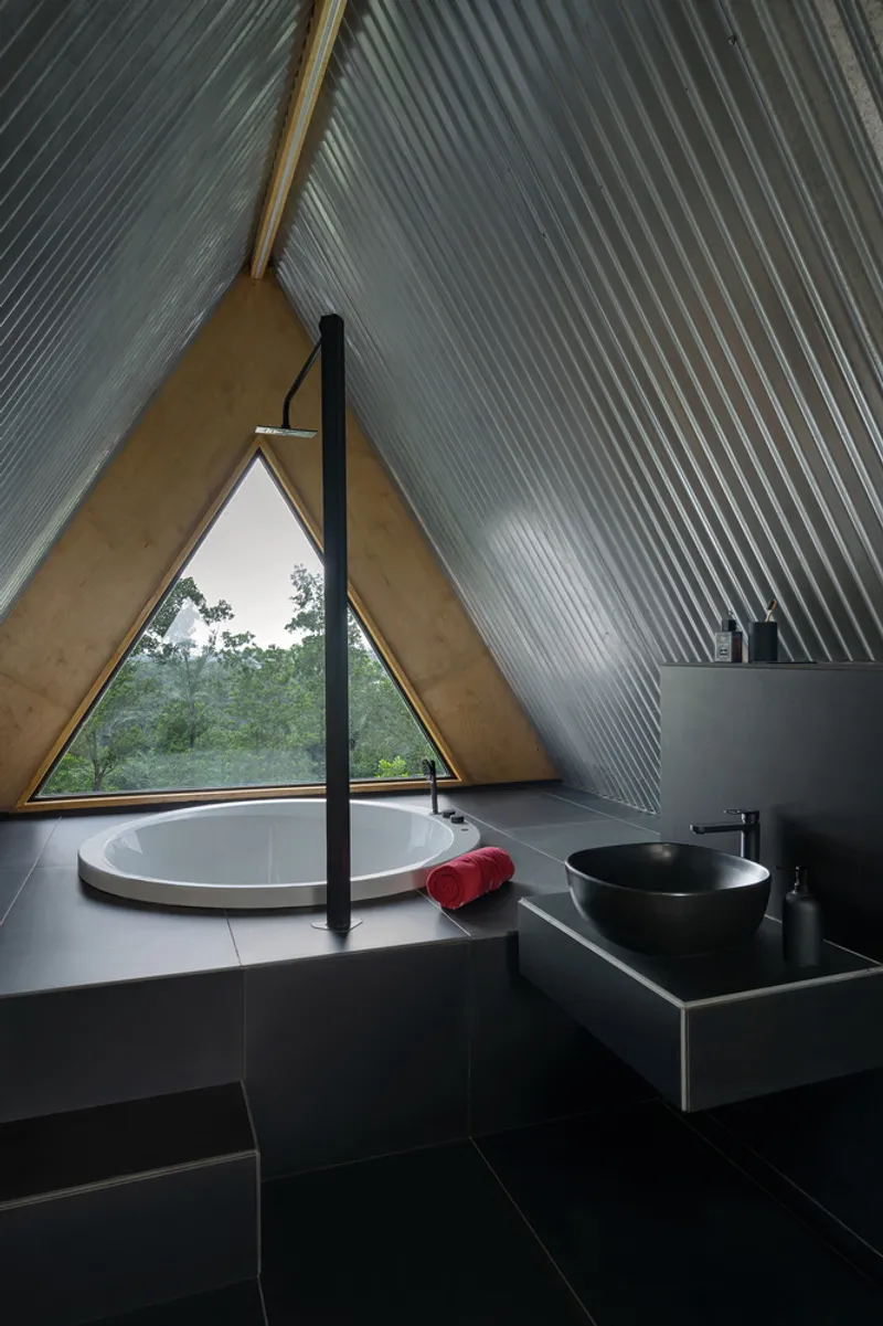 Bathroom with ribbed metal ceiling, triangular window framing forest views, and round tub with red towel