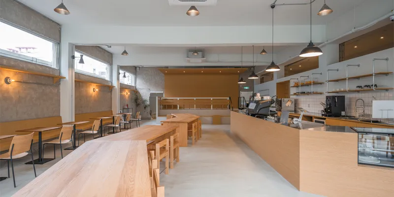 Main interior: birch plywood counter and communal table, pendant lamps, clerestory windows above exposed concrete walls, white ceiling