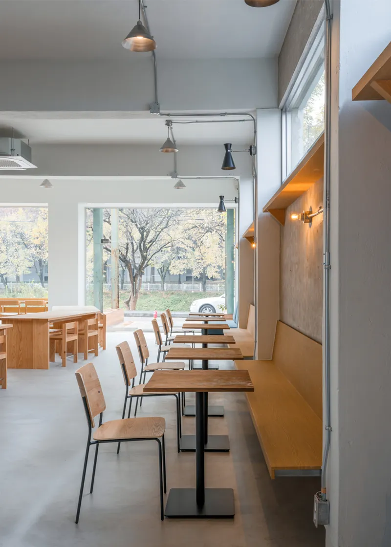 Window seating: row of chairs along the glass facade, timber shelf counter, bare winter trees visible outside, wall sconces