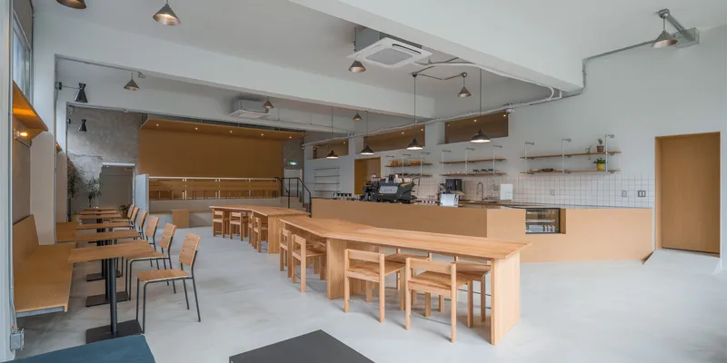 Bar counter view: birch plywood L-shaped counter, espresso machine, white tile backsplash, open shelving, stools in foreground