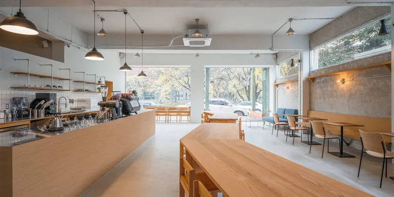 Toward the street: birch counter in foreground, full-height glass facade, autumn trees outside, exposed concrete and warm sconces