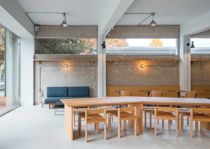 Lounge corner: exposed concrete upper walls, clerestory glass, blue sofa, birch communal table, timber ledge with warm wall lamps