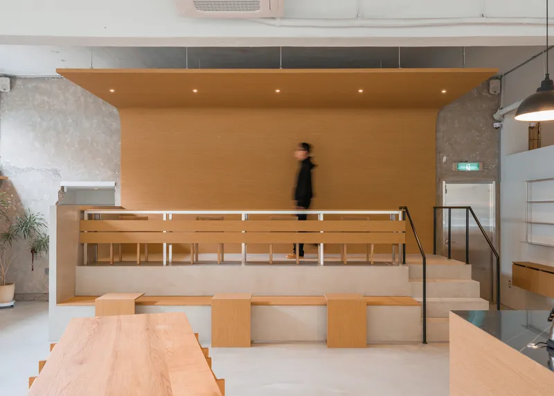 Raised platform frontal: birch plywood stepped seating under a timber ceiling, figure walking, concrete column, black railing