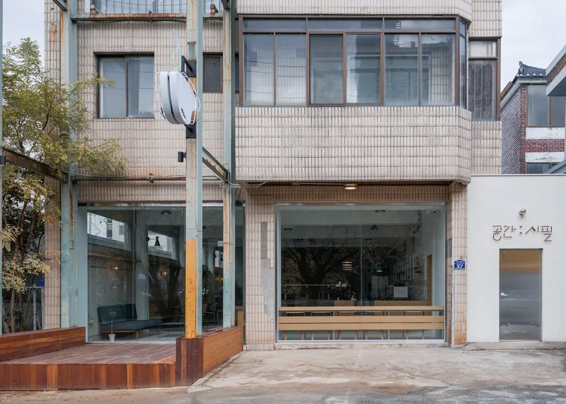 Street facade: two-storey tile-clad building, full-height glass shopfront, Korean signage, timber bench outside, quiet alley