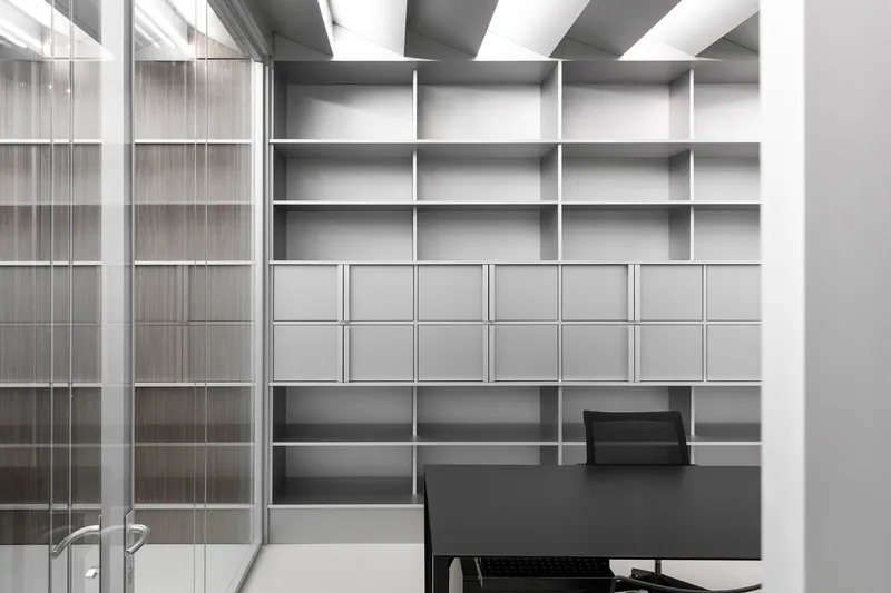 Private office: grey open shelving wall unit filling the back wall, black desk and chair, glass partition at left