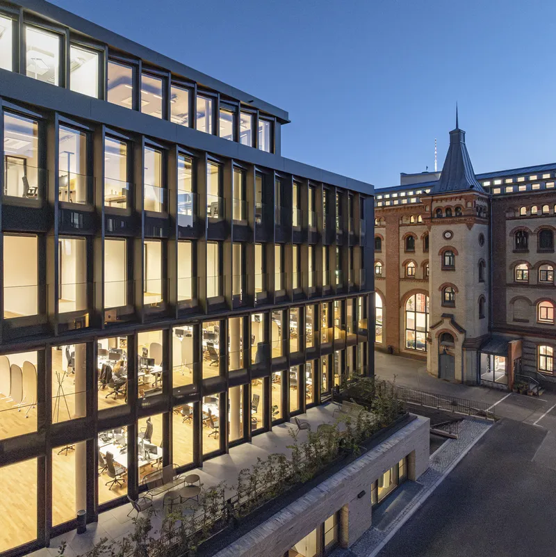 Illuminated glazed facade at dusk with planted terrace and neighboring tower in the courtyard