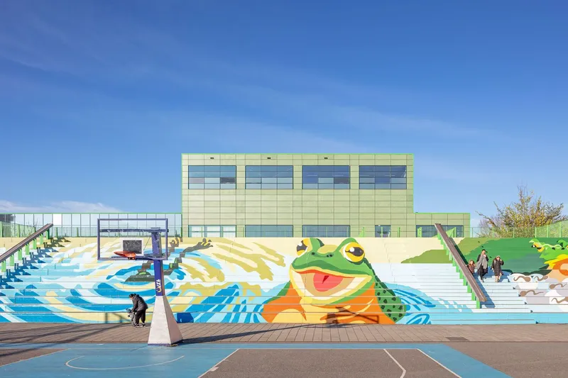 Exterior courtyard with colorful frog mural on stepped seating and basketball court in foreground