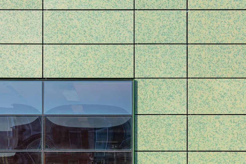 Close-up of speckled green facade panels meeting a horizontal glazed window with reflected sky