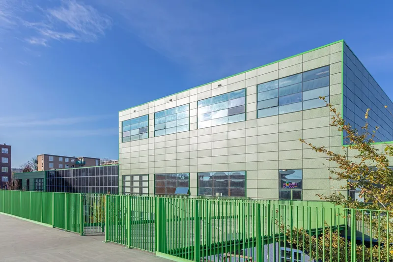 Long facade with green metal fencing and scattered trees in front of the glazed volume