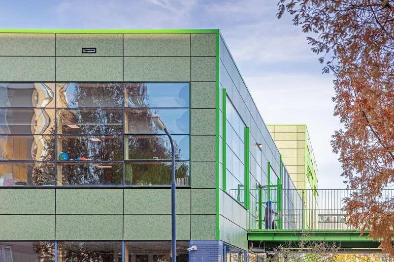 Green paneled facade with large windows reflecting bare trees and an elevated walkway connecting volumes