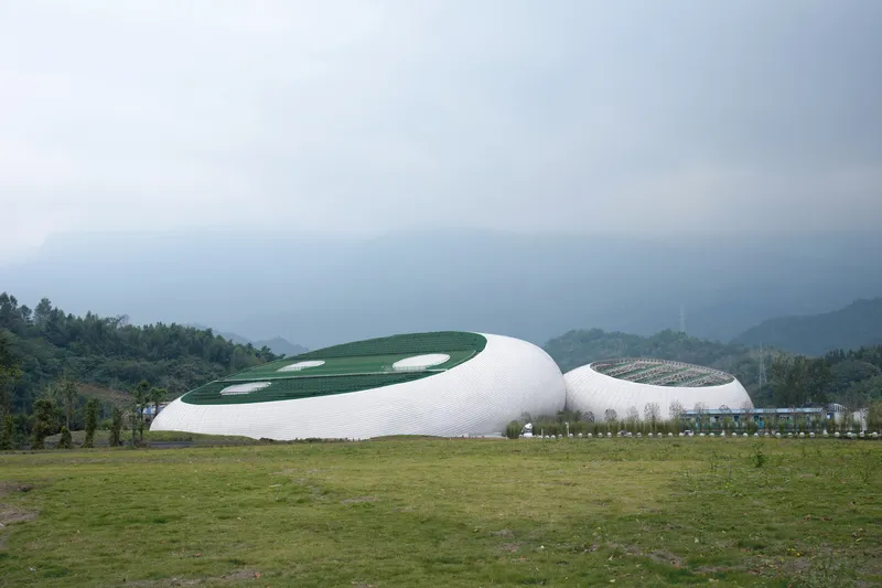 From the field: two white organic volumes with green roofs nestled against the forested hills, misty mountains beyond, overcast sky