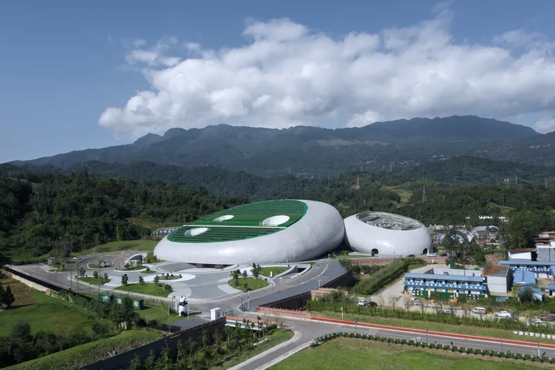 Aerial: the two cradle-shaped volumes with green roofs and circular skylights, roads and town at the perimeter, forested mountains behind
